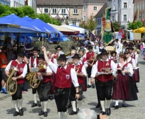 Fröhliche traditionelle Blasmusikgruppe in Trachten bei Volksfest, marschiert durch historischen Marktplatz, farbenfrohe Kleidung, Musikanten mit Instrumenten, festliche Stimmung, bayerische Kultur, Gemeinschaftsfeier, sommerliches Wetter, bayerische Festeröffnung, regionale Traditionen.