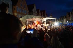 Bühne bei einem Open-Air-Konzert in einer Stadt mit überwiegend jungen Menschen, beleuchtete Gebäude im Hintergrund, abendliche Atmosphäre, Publikum genießt Live-Musik.